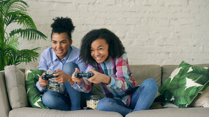 Two mixed race curly girls sitting on couch play console computer games with gamepad and have fun...