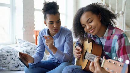 Mixed race young woman with tablet computer sitting on bed teaching her teenage sister to play acoustic guitar at home