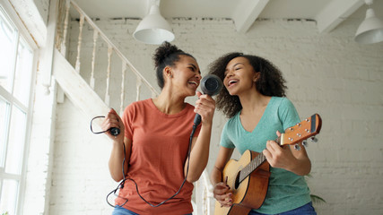 Mixed race young funny girls dance singing with hairdryer and playing acoustic guitar on a bed. Sisters having fun leisure in bedroom at home