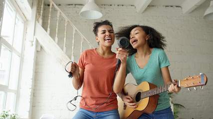Mixed race young funny girls dance singing with hairdryer and playing acoustic guitar on a bed....
