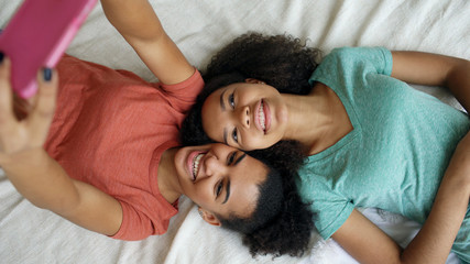 Top view of beautiful mixed race funny girlfriends making selfie portrait on bed in bedroom at home