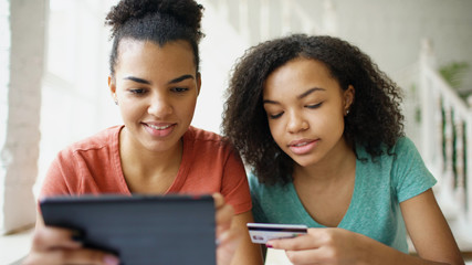 Two cheerful mixed race curly girlfriends shopping online with tablet computer and credit card at...