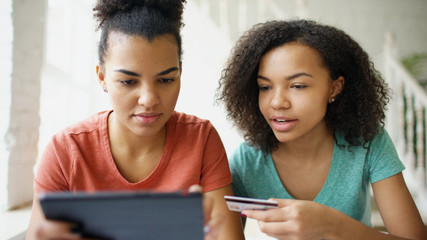 Two cheerful mixed race curly girlfriends shopping online with tablet computer and credit card at home