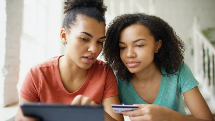 Two cheerful mixed race curly girlfriends shopping online with tablet computer and credit card at...