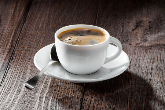 cup of coffee with nice foam close-up,  spoon and white saucer on dark wooden background
