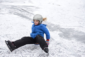 Fototapeta na wymiar Child girl falling down on ice in snowy park during winter holidays. Wearing safety helmet. Winter children activities.