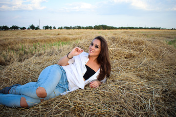 attractive smiling brunette in white shirt and jeans lies on haystack in front of field.