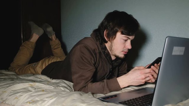 Young man typing on smartphone message, and lies on the bed in front of laptop at home.