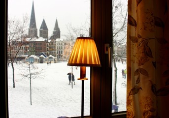 Winter square with snow view from window. Cozy interior with yellow lamp shade and curtains. Old buildings, castle and church landmark in Europe. Winter european town landscape.
