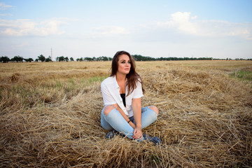 Naklejka na ściany i meble attractive smiling brunette in white shirt and jeans sitting on haystack in front of field.