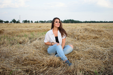 attractive smiling brunette in white shirt and jeans sitting on haystack in front of field.