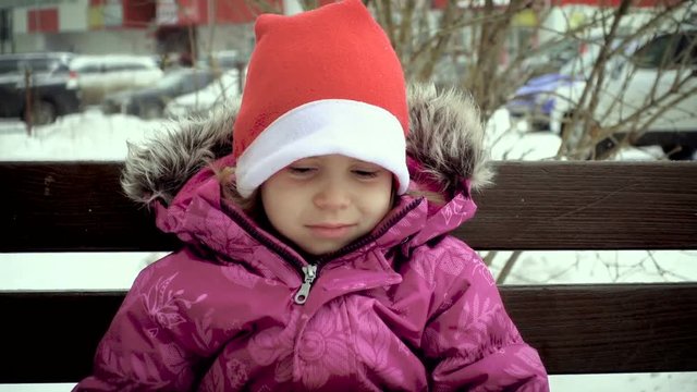 A frustrated child, a little girl, sits on a bench and waits for her mother. Mom takes pictures. Slow motion. Snowflakes fall on the girl.