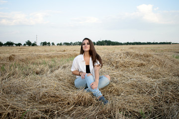 attractive smiling brunette in white shirt and jeans sitting on haystack in front of field.
