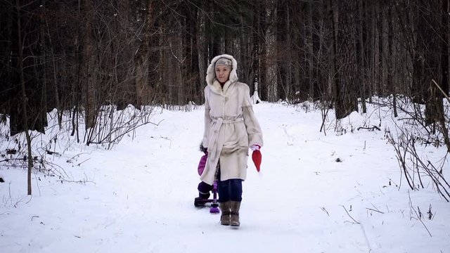Mom pulls a sled with a child. They walk in the winter forest. Daughter in a warm winter overall. A woman of European appearance in a white mink coat.