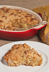 Slice of  freshly baked, homemade, warm, Apple crumb sour cream coffee cake on a white, round plate remainder in a red baking dish