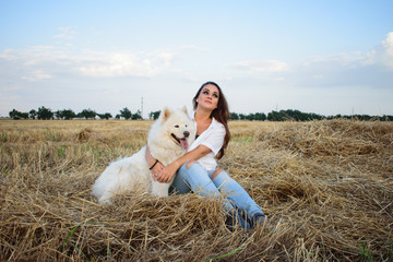 portrait of a beautiful dark-haired girl with a samoyed on the field. A young girl with her white dog is sitting on a haystack