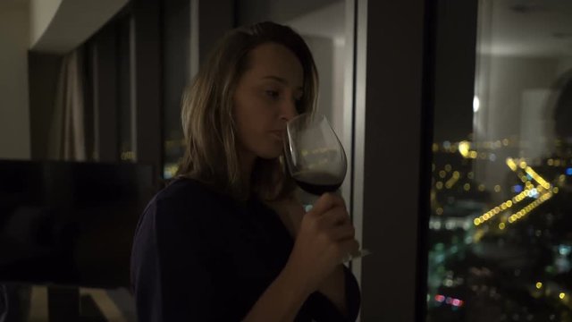 Sad, unhappy woman drinking wine admire view from window at home at night
