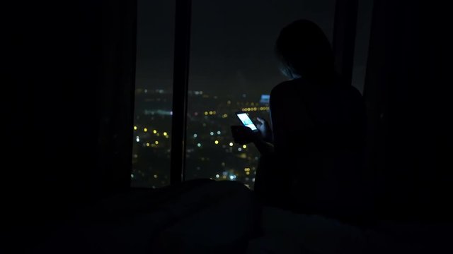 Silhouette of woman texting on smartphone on bed at night
