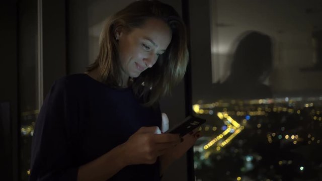 Young woman texting on smartphone standing by window at home at night

