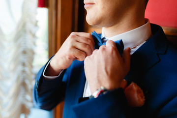 Closeup of a gentleman in an expensive suit wearing a blue tie, straightens his bow tie.