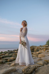 Elegant young woman in white long dress on sea background. Bride with flowers at sunset, sunrise,dawn