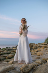 Elegant young woman in white long dress on sea background. Bride with flowers at sunset, sunrise,dawn