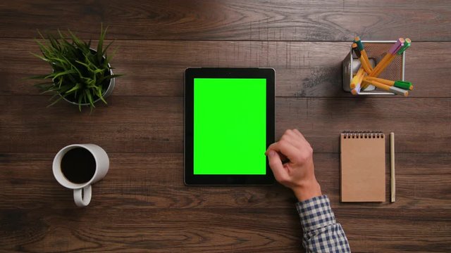 A man's finger scrolling on the green touchscreen. The i-Pad is on the brown table. View from the top. Close-up.