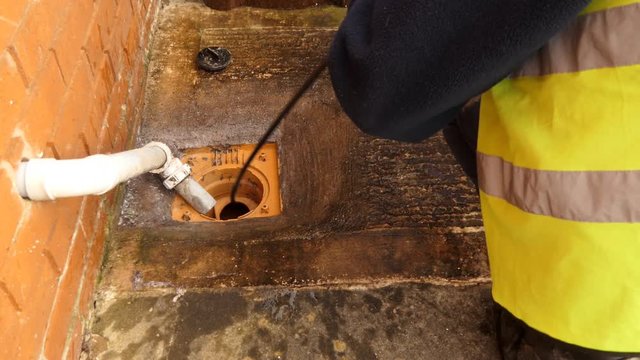 Plumber wearing bright yellow safety vest working by residential house outdoors pulling out drain rod sewer snake with camera and light from sewage pipe after finding,cleaning blockage in sewer line