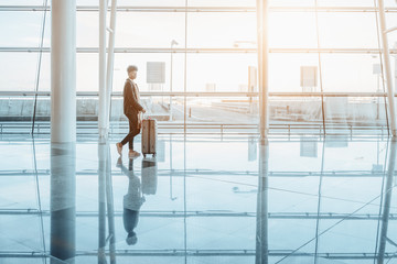 Black girl in warm clothes and with massive bag with wheels is standing alone inside of bright interior of modern airport terminal on reflective floor near contemporary glass facade