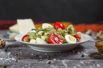 pasta from green spinach with mozzarella, greens and French mustard, quail eggs and cherry tomatoes on a plate, bell pepper and large sea salt on a gray wooden table on a dark background
