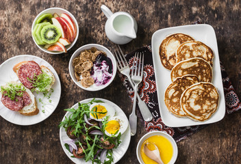 Breakfast table - greek yogurt with whole grain cereals and berry sauce, pancakes, arugula, cherry tomatoes, boiled eggs salad, kiwi, apples fruit, salami and cream cheese sandwiches 