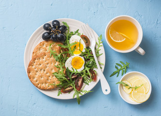 Breakfast plate - cracker, arugula, cherry tomatoes, boiled egg salad and green tea with lemon on blue background, top view