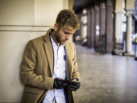 Handsome Trendy Blond Man Wearing Dark Coat Standing And Looking Down At A Cell Phone That He Is Holding, Outdoor In European City Setting With Elegant Old Historic Building Behind