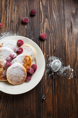 syrniki with powdered sugar on a white plate, sieve and frozen berries on a dark wooden table
