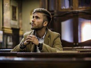 Young blond elegant man sitting in church, on wooden bench, thinking and praying, looking around