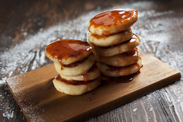 syrniki with powdered sugar on a white plate, a sieve and caramel topping on a dark wooden table