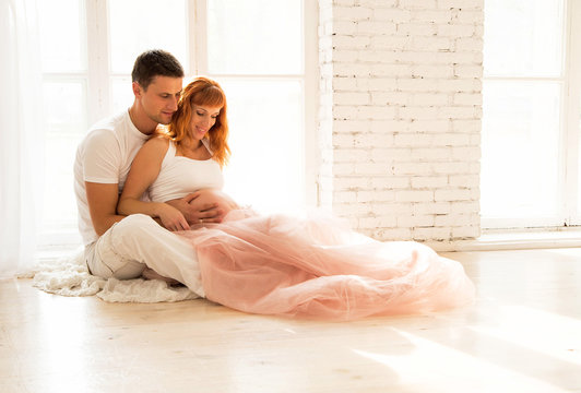pregnant woman in a tulle skirt and her husband are sitting on the floor near a large window