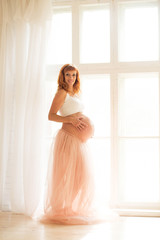 A pregnant woman in a white top and a tulle skirt stands near a large window