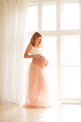 A pregnant woman in a white top and a tulle skirt stands near a large window