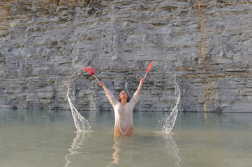 A young beautiful hippie girl is having fun with two large red hearts in the water. She sings and shouts  out loud while splashing water around her.
