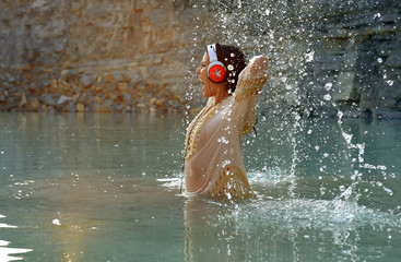 A young beautiful girl with headphones on her head is having fun with music in the water. She sings and  shouts out loud while splashing water around her.