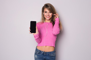 Smiling blonde woman in pink blouse showing blank smartphone screen and listening to music in headphones isolated on a gray background