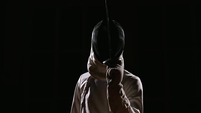 A man dressed for fencing wears a mask and is preparing a rapier. Close-up. The position to start sparring
