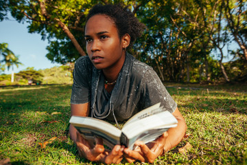 Beautiful young woman with afro hairstyle and book in her hands lying on the grass in the park on...