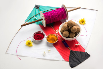 happy Makar Sankranti Festival - Tilgul or Til ladoo in a bowl or plate with haldi kumkum and flowers with Fikri /Reel/Chakri /Spool with colourful thread or manjha and kite over plain background
