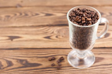 Lot of fried coffee beans in transparent glass for mulled wine with handle and leg beside scattered grains on old wooden rustic brown planks