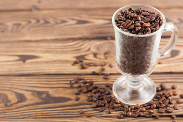 Lot of fried coffee beans in transparent glass for mulled wine with handle and leg beside scattered many grains on old wooden rustic brown planks