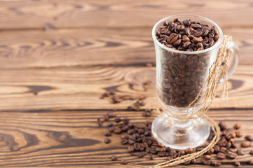 Lot of fried coffee beans in transparent glass for mulled wine with brown rope beside scattered many grains on old wooden rustic brown planks