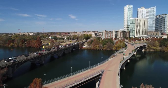 A slow forward rising aerial establishing shot (DX) of the Austin city skyline with the Pfluger Pedestrian Bridge over the Colorado River in the foreground on a late sunny Autumn day.  	