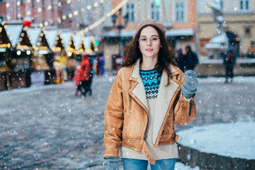 Portrait of woman in woolen sweater, beret and leather jacket enjoying winter moments. Outdoor photo of long-haired inspiring lady having fun in snowy evening over old town square with Christmas fair.